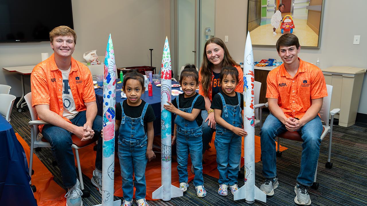 three students in orange shirts pose for a photo with three girls in overalls holding painted rockets