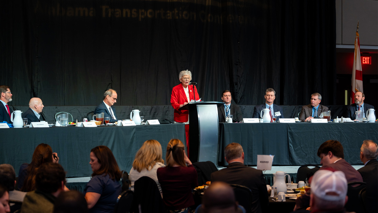 Gov. Kay Ivey speaks at a podium during the Alabama Transportation Conference, with Auburn’s Larry Rilett and Alabama Department of Transportation officials seated nearby.
