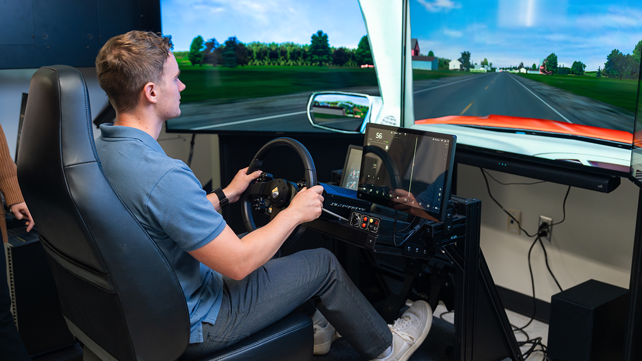 A student uses a driving simulator in a laboratory, seated at a steering wheel with multiple screens displaying a simulated roadway.