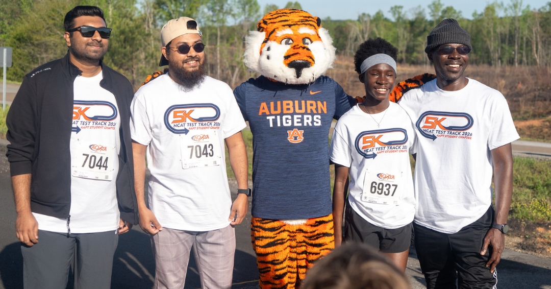 Auburn University mascot Aubie poses with participants and AAPT student chapter members at the NCAT Test Track 5K, with runners wearing race bibs and event t-shirts on the track.