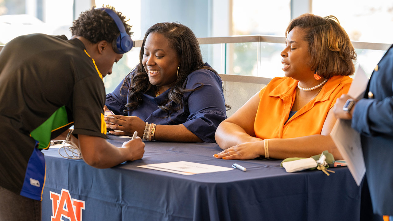 A student fills out a form on a blue table in front of two women