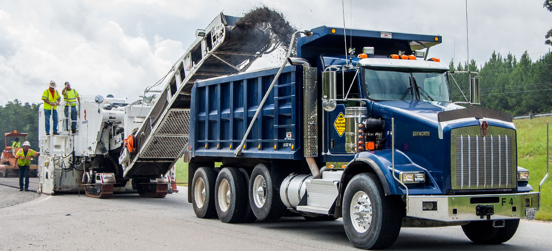 Milling machine loads recycled asphalt into dump truck while workers overlook process